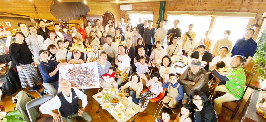 老若男女が集まった集合写真 カタツムリのイラストが描かれた旗を数人でカメラに向けて広げていたり机には料理があったりと楽しげな雰囲気