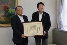 みやぎ千本桜愛護会の表敬訪問の写真です。代表者の男性と市長が一緒に、額に入った賞状を持っています。