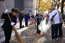 歩道にあるたくさんの落ち葉をほうきで掃くボランティアの人たち