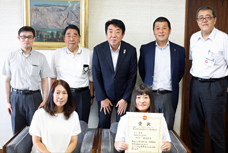 全日本高校ボウリング選手権大会優勝者の表敬訪問の写真です。前列左に女子生徒の母親が、右側に女子生徒が座っていて、女子生徒は賞状を持っています。後列には高校の職員を含めた5人の男性がいて、真ん中に市長がいます。