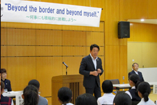 中学生海外研修事業到着式で市長がお話をしている写真です。ステージ上には、スローガンである「Beyond the border and beyond myself.～何事にも積極的に挑戦しよう～」という言葉が書かれた幕が張ってあります。