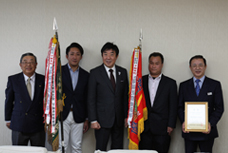 第64回県町内対抗軟式野球大会優勝に伴う表敬訪問で、優勝旗を持った代表と市長が並んでいる写真です。